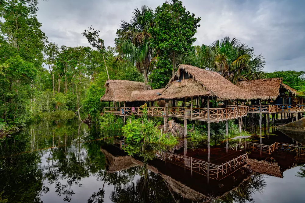 Linda vista da floresta amazônica em um dia ensolarado perfeito, com cabanas de madeira sobre o rio refletindo a vegetação exuberante e o céu claro.