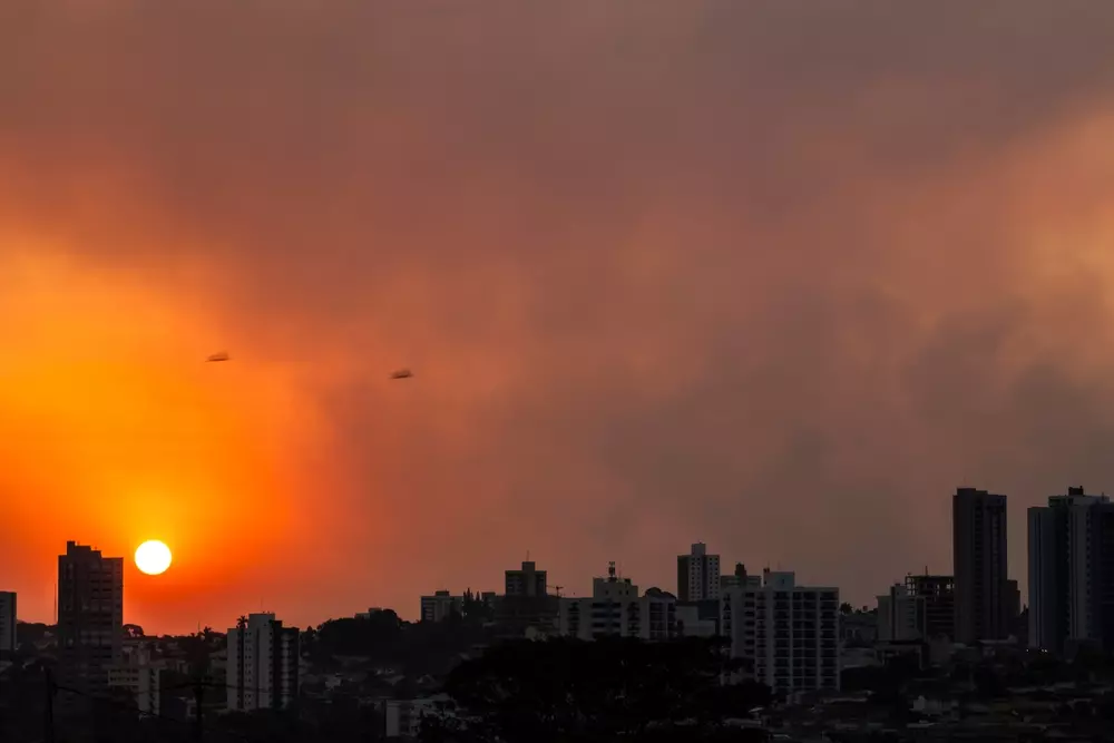 Cidade urbana durante o pôr do sol, com fumaça e céu com tons alaranjados e escuros, ilustrando o fenômeno das ilhas de calor, desigualdade e planejamento urbano.