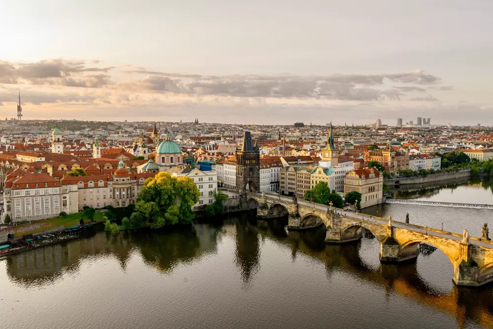 Vista panorâmica da cidade de Praga com o rio Vltava ao centro, destacando a Ponte Charles, edifícios históricos e o pôr do sol ao fundo, ilustrando a beleza arquitetônica e cultural da capital tcheca.