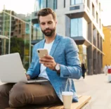 Homem de negócios usando celular e trabalhando com laptop ao ar livre em cidade moderna, representando as melhores cidades para trabalho remoto.
