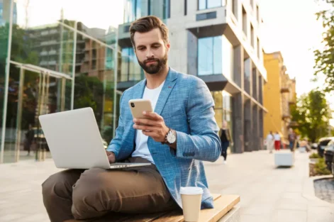 Homem de negócios usando celular e trabalhando com laptop ao ar livre em cidade moderna, representando as melhores cidades para trabalho remoto.