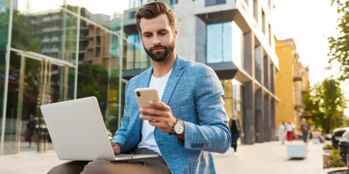Homem de negócios usando celular e trabalhando com laptop ao ar livre em cidade moderna, representando as melhores cidades para trabalho remoto.
