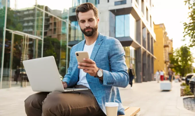 Homem de negócios usando celular e trabalhando com laptop ao ar livre em cidade moderna, representando as melhores cidades para trabalho remoto.
