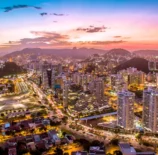 Vista panorâmica da cidade do Rio de Janeiro ao entardecer, com luzes iluminando os prédios, montanhas ao fundo e o céu colorido ao pôr do sol.