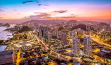 Vista panorâmica da cidade do Rio de Janeiro ao entardecer, com luzes iluminando os prédios, montanhas ao fundo e o céu colorido ao pôr do sol.