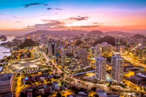Vista panorâmica da cidade do Rio de Janeiro ao entardecer, com luzes iluminando os prédios, montanhas ao fundo e o céu colorido ao pôr do sol.