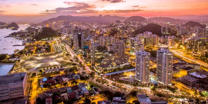 Vista panorâmica da cidade do Rio de Janeiro ao entardecer, com luzes iluminando os prédios, montanhas ao fundo e o céu colorido ao pôr do sol.