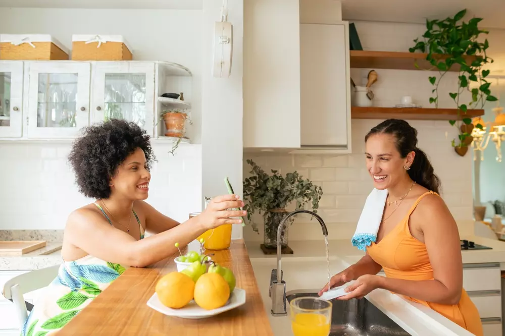 Duas mulheres felizes conversando na cozinha moderna, uma segurando um copo de suco e a outra lavando a louça, ambiente acolhedor e bem iluminado