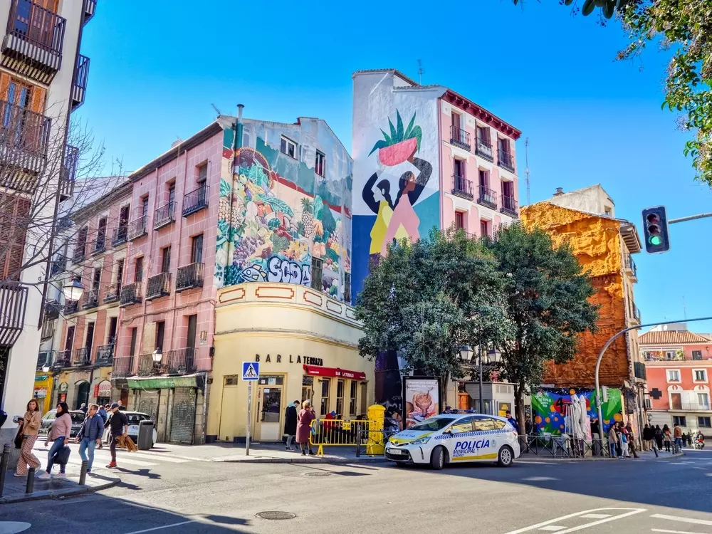 Prédio de parede decorada com mural artístico, árvores, carros da polícia, e várias pessoas caminhando na cidade em um dia ensolarado.