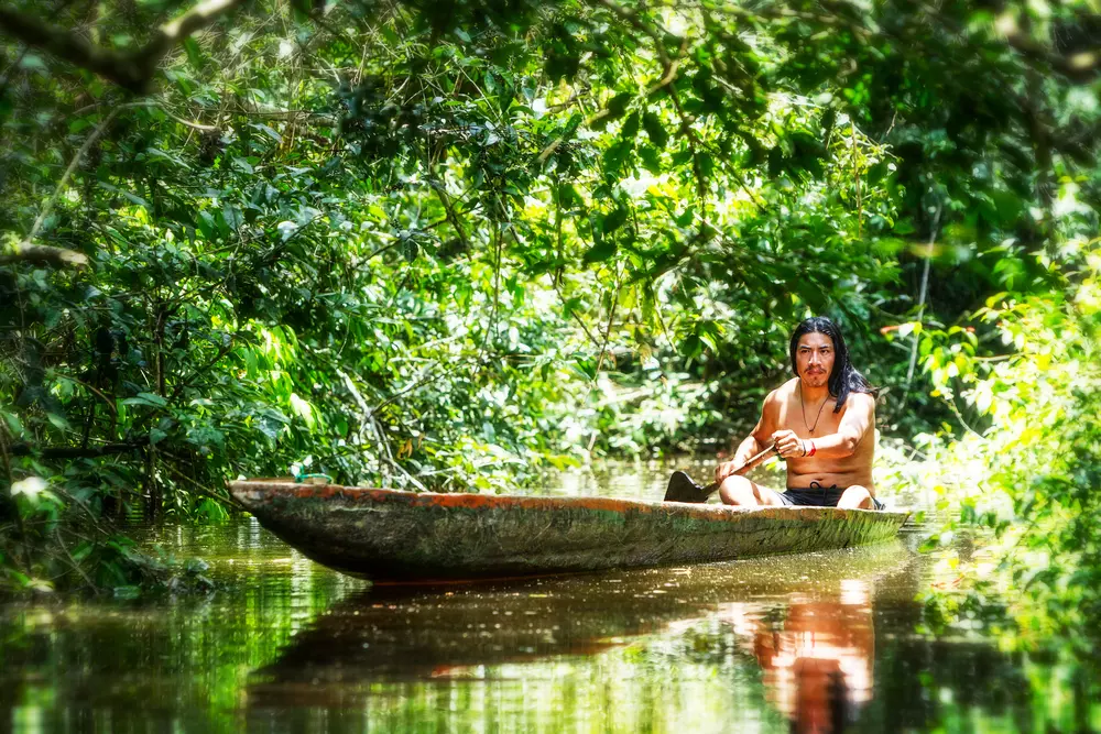 Homem indígena navegando por um rio na floresta, simbolizando a conexão com a natureza e a luta por justiça climática, preservando ecossistemas e combatendo a mudança climática.