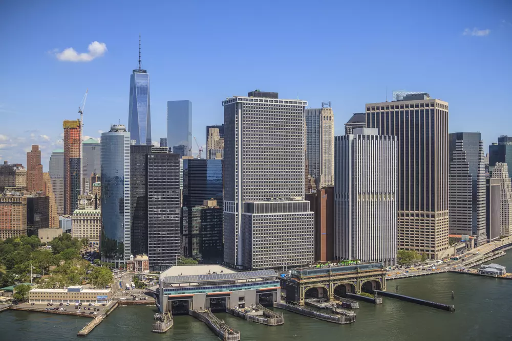 Foto da skyline de Manhattan, Nova York, com edifícios altos e o rio em primeiro plano, sob céu azul claro.