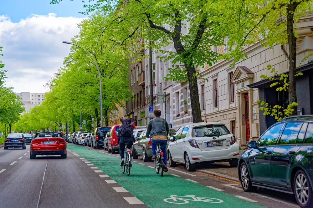 Cena de uma rua de uma cidade com árvores verdes, carros estacionados e ciclistas usando a ciclofaixa, refletindo o valor de uma cidade sustentável e de alta qualidade de vida. Ilustrando parte da matéria sobre reputação que molda o valor de marca de um país
