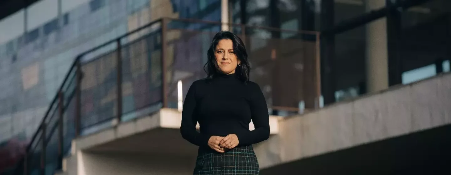Fotografia de Ana Carolina Vaz em frente a uma construção moderna, vestindo blusa preta e saia de xadrez, com expressão confiante e cenário urbano ao fundo.