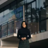 Fotografia de Ana Carolina Vaz em frente a uma construção moderna, vestindo blusa preta e saia de xadrez, com expressão confiante e cenário urbano ao fundo.