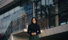 Fotografia de Ana Carolina Vaz em frente a uma construção moderna, vestindo blusa preta e saia de xadrez, com expressão confiante e cenário urbano ao fundo.