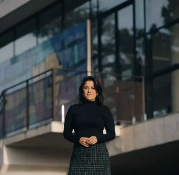 Fotografia de Ana Carolina Vaz em frente a uma construção moderna, vestindo blusa preta e saia de xadrez, com expressão confiante e cenário urbano ao fundo.