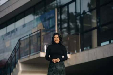 Fotografia de Ana Carolina Vaz em frente a uma construção moderna, vestindo blusa preta e saia de xadrez, com expressão confiante e cenário urbano ao fundo.
