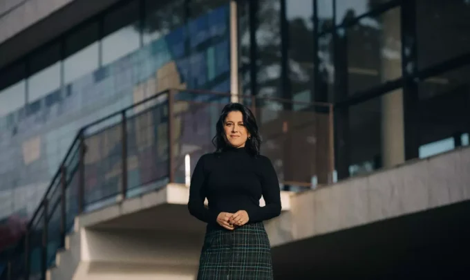 Fotografia de Ana Carolina Vaz em frente a uma construção moderna, vestindo blusa preta e saia de xadrez, com expressão confiante e cenário urbano ao fundo.