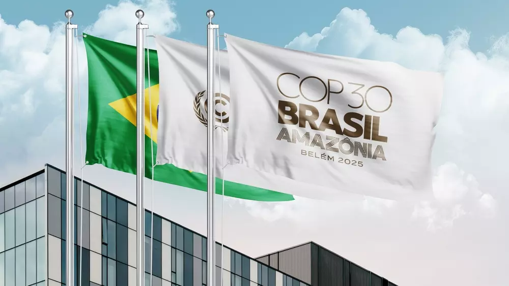 Bandeiras do Brasil e da COP Brasil Amazônia tremulam em frente a um prédio moderno durante o evento COP30 em Belém, onde foram estabelecidos os primeiros padrões globais para edificações sustentáveis e resilientes.