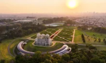 Pôr do sol no horizonte de Curitiba com vista aérea de um bairro sustentável, destacando jardins bem planejados e uma estufa moderna na paisagem