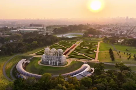 Pôr do sol no horizonte de Curitiba com vista aérea de um bairro sustentável, destacando jardins bem planejados e uma estufa moderna na paisagem