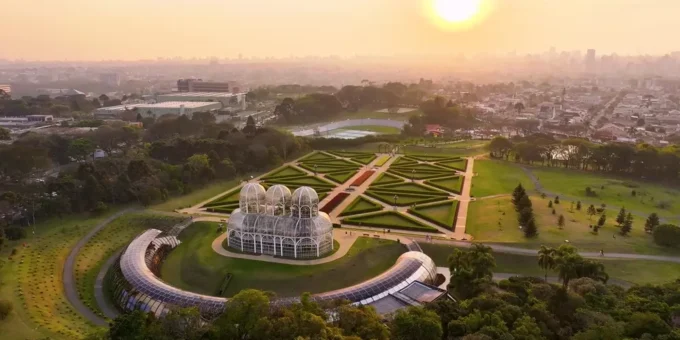 Pôr do sol no horizonte de Curitiba com vista aérea de um bairro sustentável, destacando jardins bem planejados e uma estufa moderna na paisagem