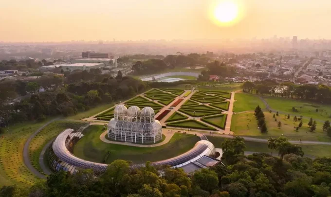 Pôr do sol no horizonte de Curitiba com vista aérea de um bairro sustentável, destacando jardins bem planejados e uma estufa moderna na paisagem
