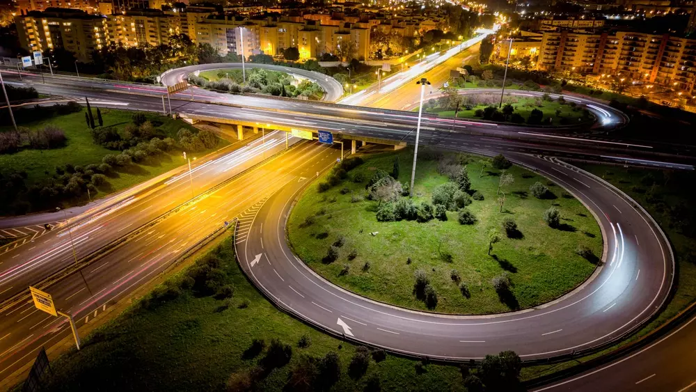Elementos geométricos e sinalização em uma rotatória iluminada à noite, com curvas, sinais de trânsito e luzes urbanas destacando a organização viária.