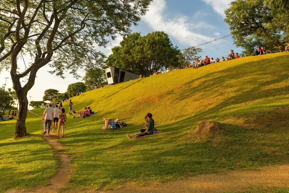 Pessoas aproveitando um dia ao ar livre em um parque com gramado, árvores e uma colina, com várias atividades sociais ao entardecer.
