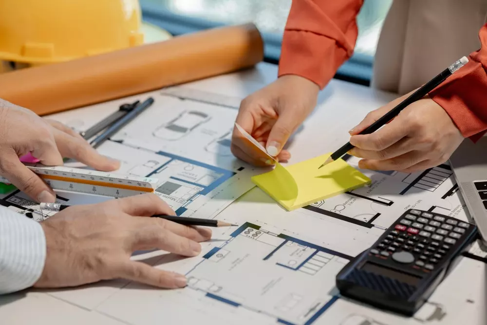Profissionais analisando e desenhando plantas de arquitetura com ferramentas de precisão, como régua, escalímetro, post-its e calculadora