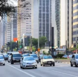 uma rua movimentada em uma cidade moderna, com veículos diversos, árvores e prédios altos ao fundo, evidenciando o streetscape