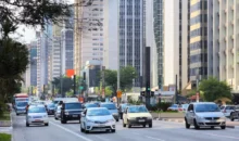 uma rua movimentada em uma cidade moderna, com veículos diversos, árvores e prédios altos ao fundo, evidenciando o streetscape