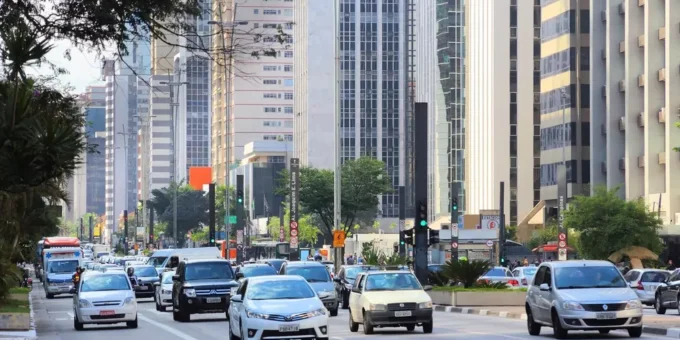 uma rua movimentada em uma cidade moderna, com veículos diversos, árvores e prédios altos ao fundo, evidenciando o streetscape