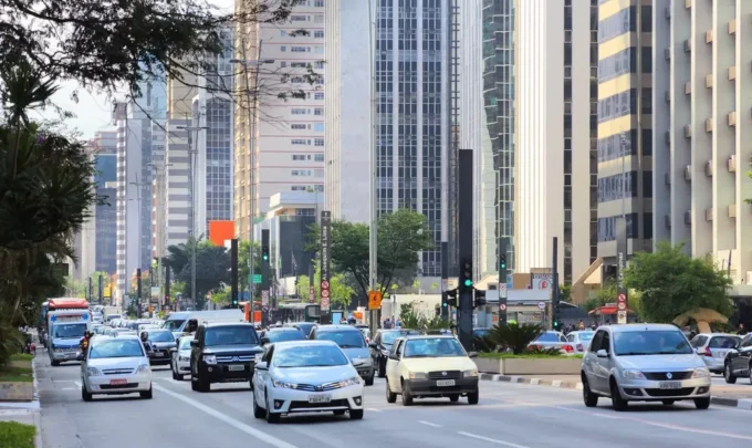 uma rua movimentada em uma cidade moderna, com veículos diversos, árvores e prédios altos ao fundo, evidenciando o streetscape