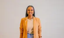 Audrey Carolini, arquiteta, urbanista e designer, posando com sorriso e vestindo blazer bege ao lado de parede neutra