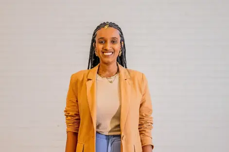 Audrey Carolini, arquiteta, urbanista e designer, posando com sorriso e vestindo blazer bege ao lado de parede neutra