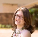 Retrato de Karla Zambrano sorridente ao ar livre, com fundo desfocado, usando óculos e vestido floral.