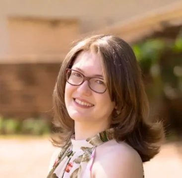 Retrato de Karla Zambrano sorridente ao ar livre, com fundo desfocado, usando óculos e vestido floral.