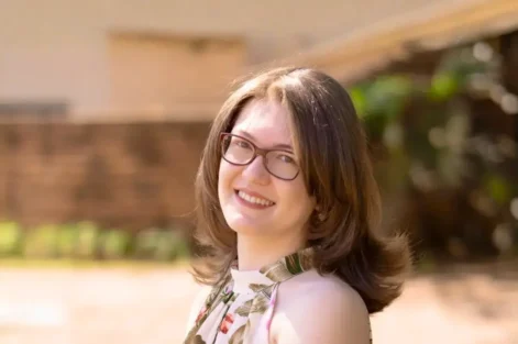 Retrato de Karla Zambrano sorridente ao ar livre, com fundo desfocado, usando óculos e vestido floral.