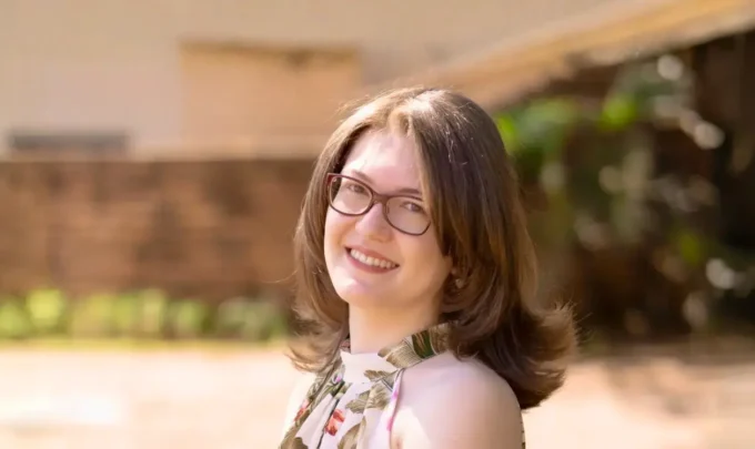 Retrato de Karla Zambrano sorridente ao ar livre, com fundo desfocado, usando óculos e vestido floral.