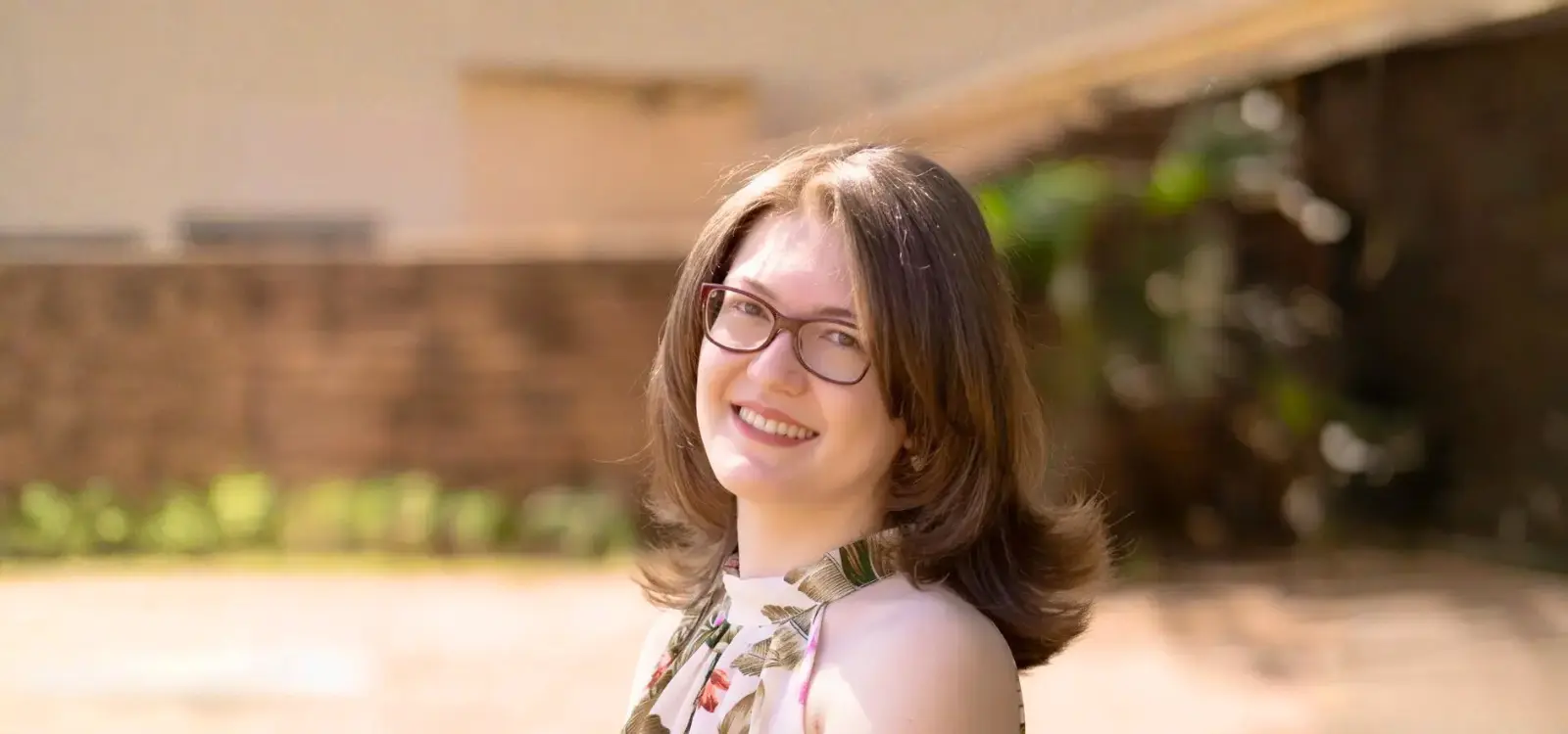 Retrato de Karla Zambrano sorridente ao ar livre, com fundo desfocado, usando óculos e vestido floral.