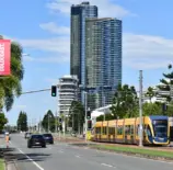 Um cenário urbano com um trem elétrico amarelo em uma via de transporte, altos prédios ao fundo e árvores, destacando a mobilidade sustentável e o uso de carros elétricos na cidade.