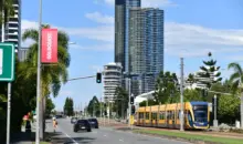 Um cenário urbano com um trem elétrico amarelo em uma via de transporte, altos prédios ao fundo e árvores, destacando a mobilidade sustentável e o uso de carros elétricos na cidade.