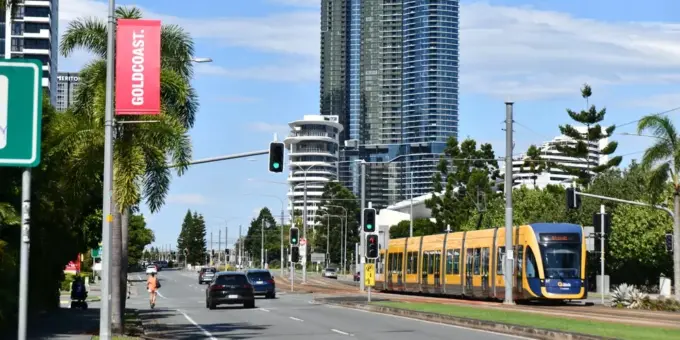 Um cenário urbano com um trem elétrico amarelo em uma via de transporte, altos prédios ao fundo e árvores, destacando a mobilidade sustentável e o uso de carros elétricos na cidade.