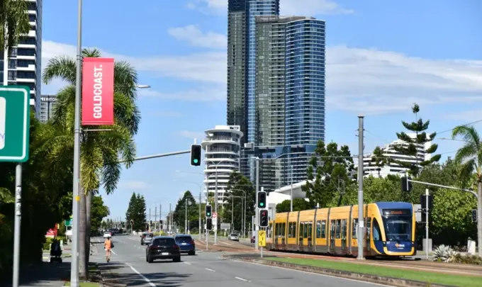 Um cenário urbano com um trem elétrico amarelo em uma via de transporte, altos prédios ao fundo e árvores, destacando a mobilidade sustentável e o uso de carros elétricos na cidade.