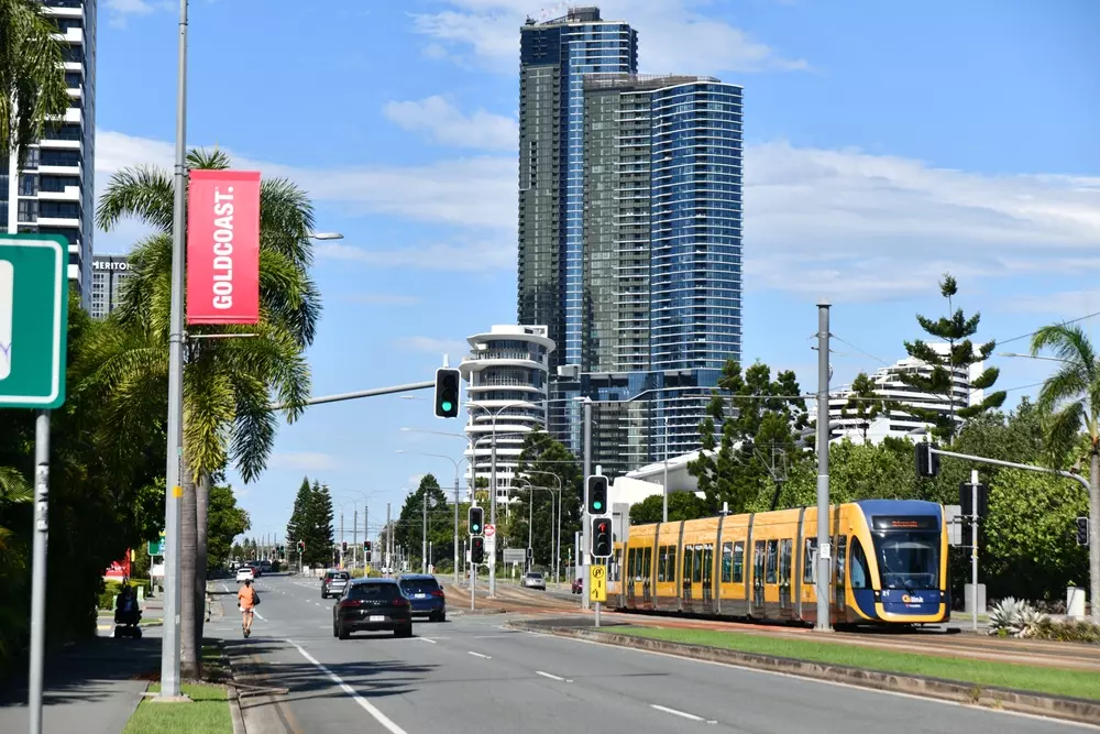 Um cenário urbano com um trem elétrico amarelo em uma via de transporte, altos prédios ao fundo e árvores, destacando a mobilidade sustentável e o uso de carros elétricos na cidade.