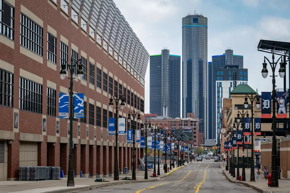 Centro de Detroit com banners do Comerica Park e Ford Field, destaque para arranha-céus no horizonte, ruas tranquilas e elementos urbanos característicos da região.