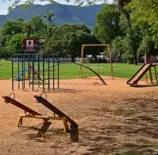 Parque com equipamentos públicos de lazer, incluindo brinquedos e estruturas de ginástica, em área verde com árvores e montanhas ao fundo