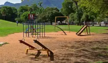Parque com equipamentos públicos de lazer, incluindo brinquedos e estruturas de ginástica, em área verde com árvores e montanhas ao fundo