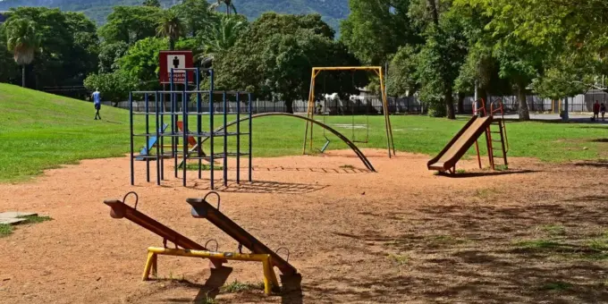 Parque com equipamentos públicos de lazer, incluindo brinquedos e estruturas de ginástica, em área verde com árvores e montanhas ao fundo